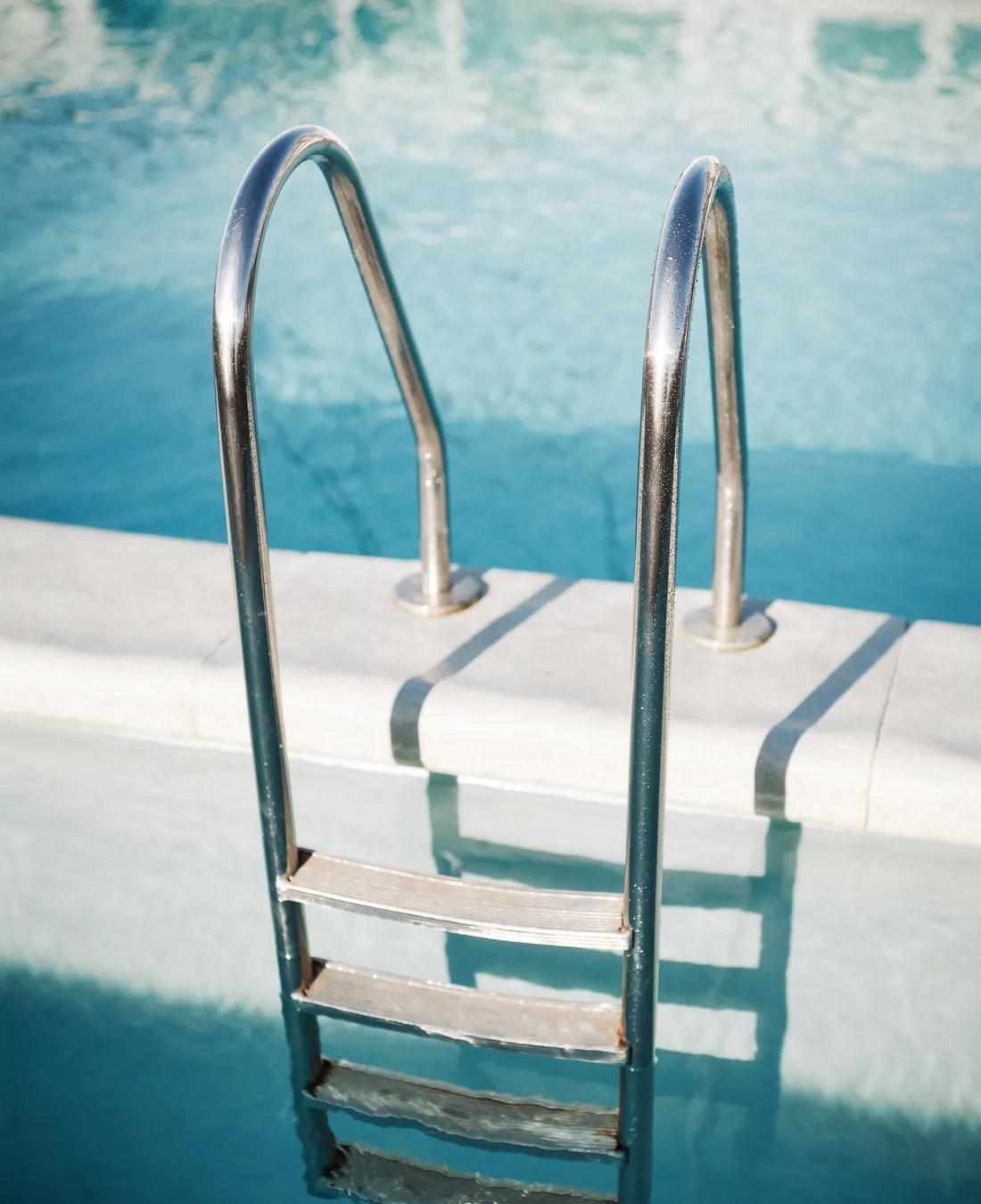 A stainless steel pool ladder with two handrails rising out of clear blue water, seen close from the deck, with the pale liner visible below the waterline.