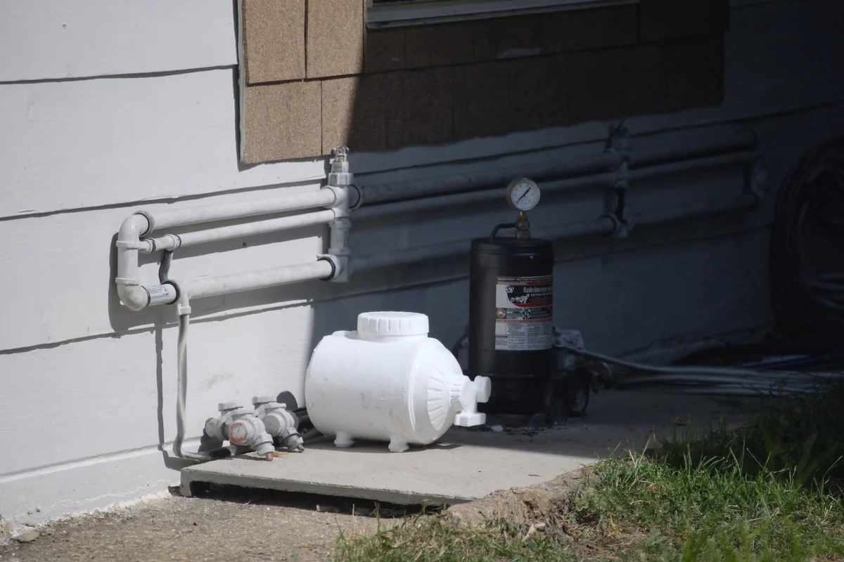 A residential pool equipment pad on a concrete slab beside a house wall, with a white pump, a tall sand filter carrying a pressure gauge, grey PVC pipework and valves, and a coiled garden hose.