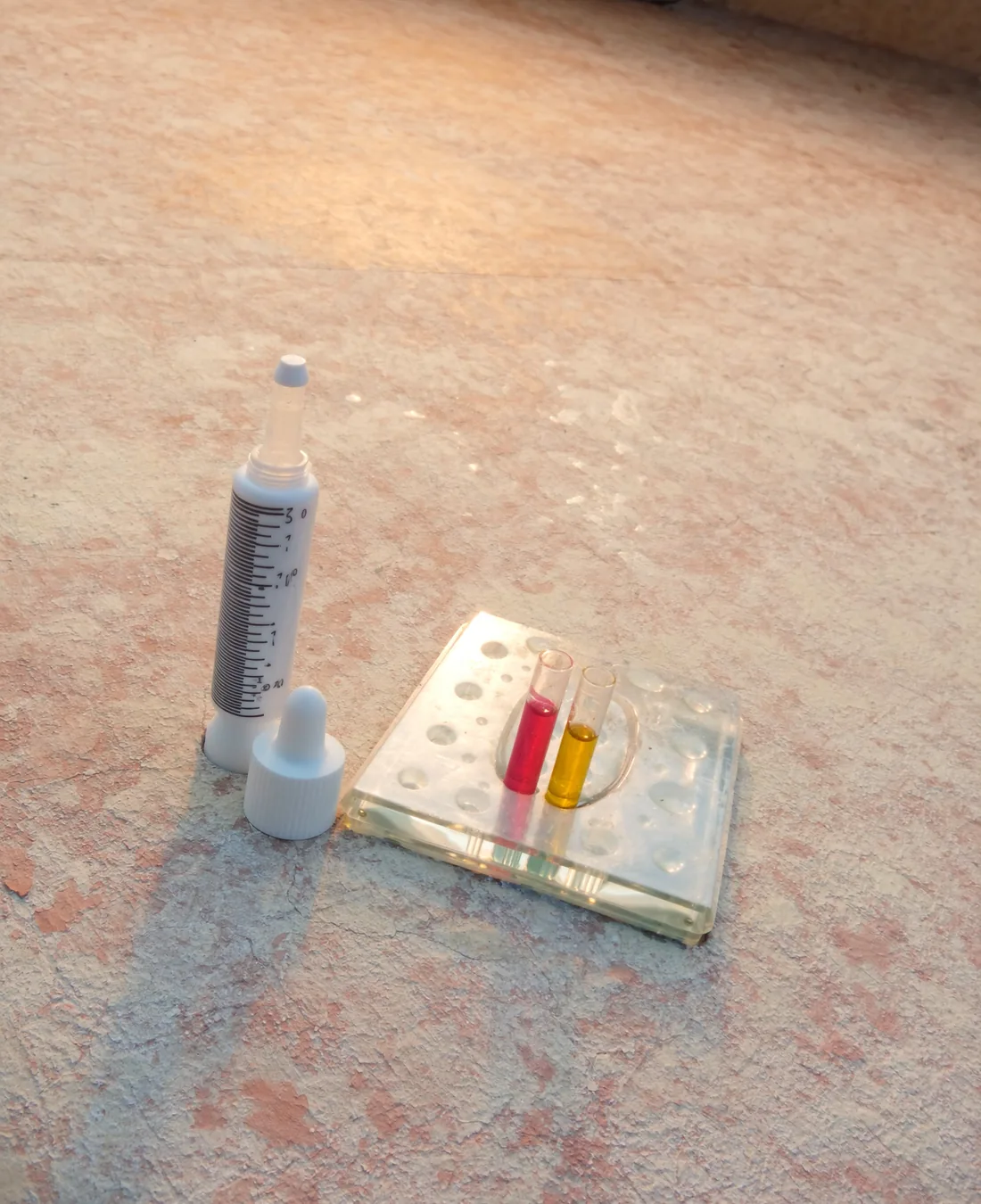 A pool water test kit open on a wet concrete deck, with a moulded comparator block holding two small vials of tinted water, one pink and one yellow, and a dropper bottle of reagent beside it.